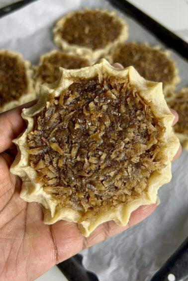 A hand holding a freshly baked gizzada with a close-up of the pinched crust and sticky coconut filling, with more gizzadas blurred in the background.