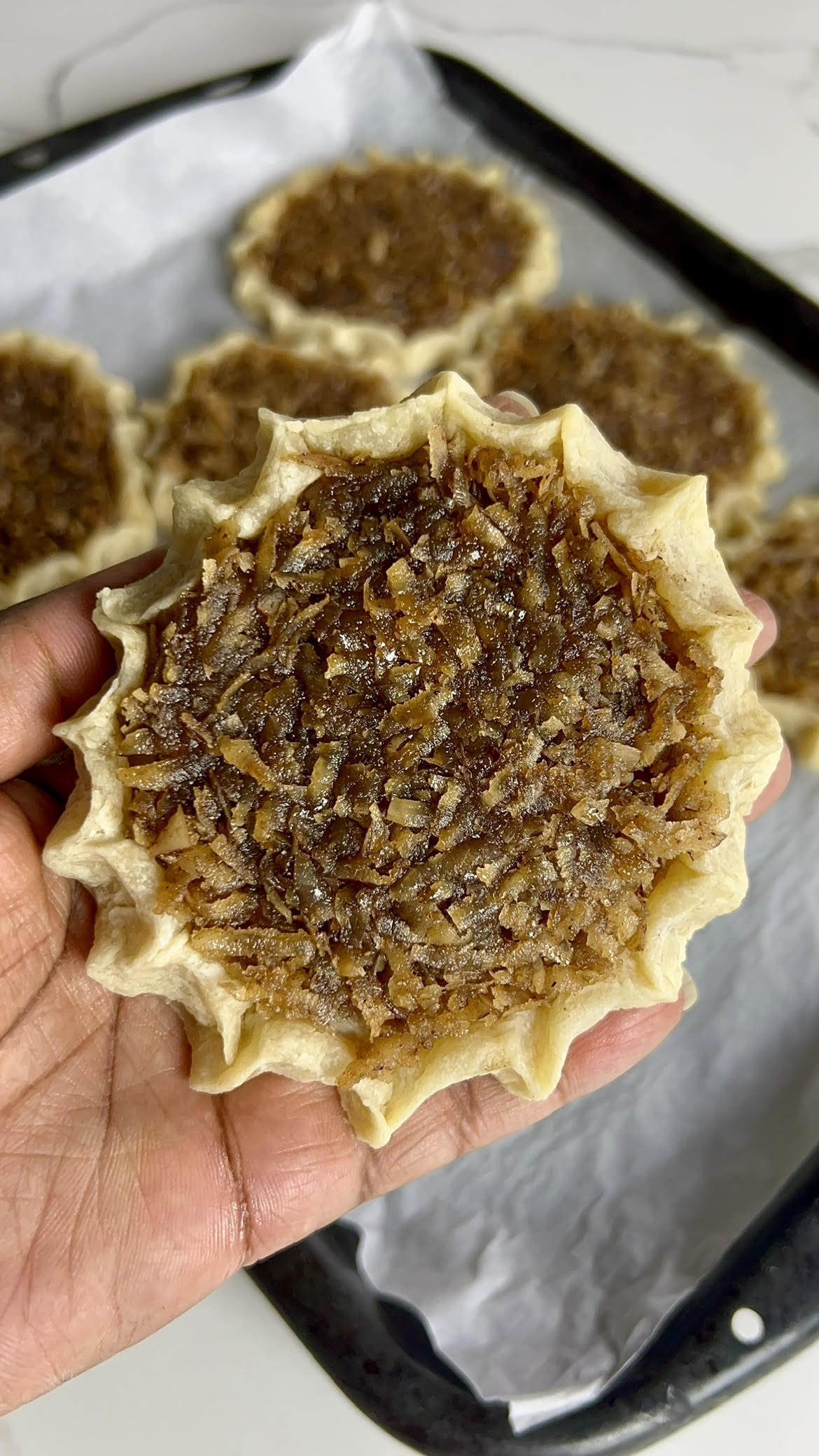A hand holding a freshly baked gizzada with a close-up of the pinched crust and sticky coconut filling, with more gizzadas blurred in the background.