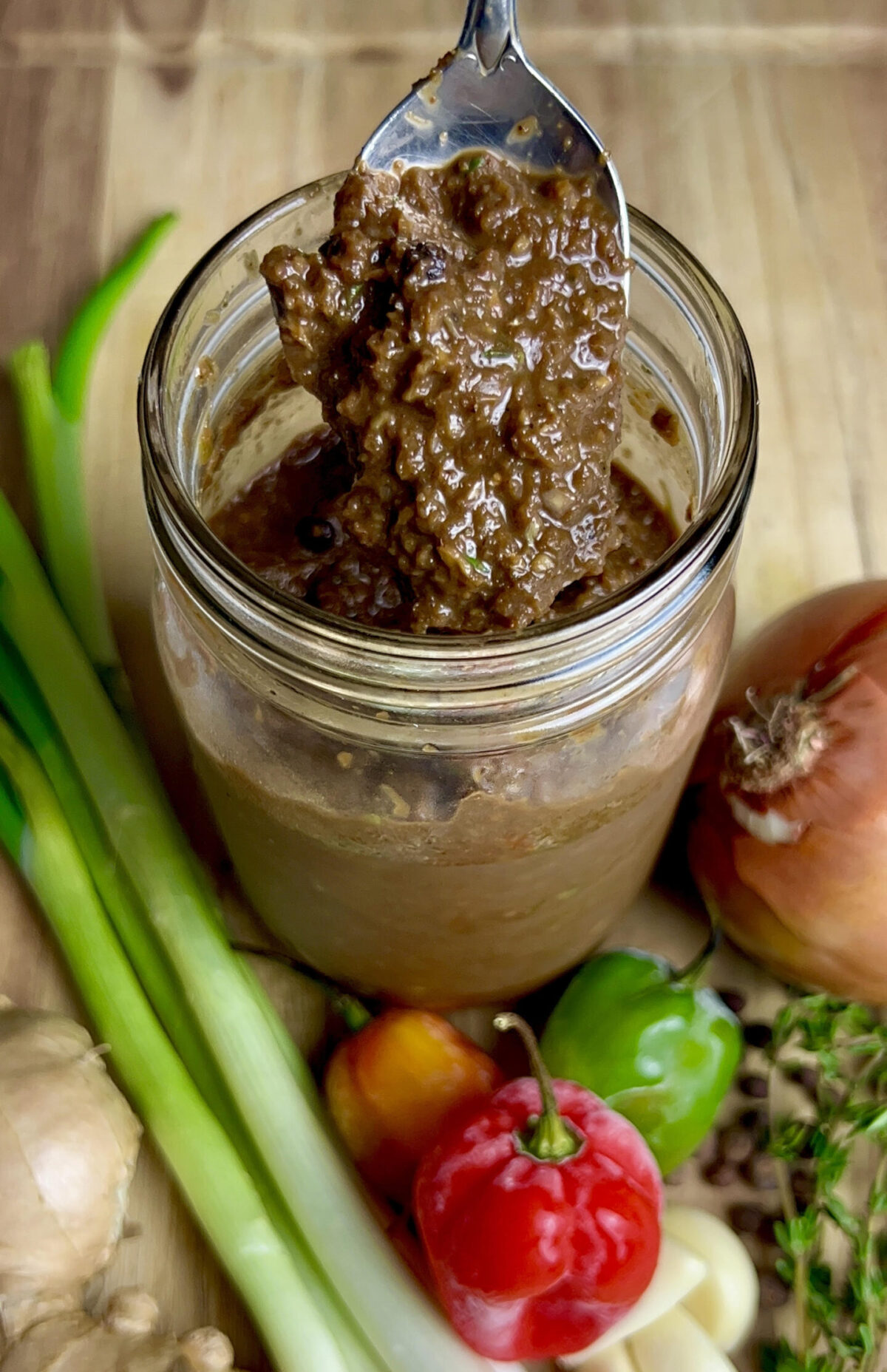 A jar of homemade jerk marinade surrounded by fresh ingredients like scallions, peppers, garlic, ginger, and thyme on a cutting board.