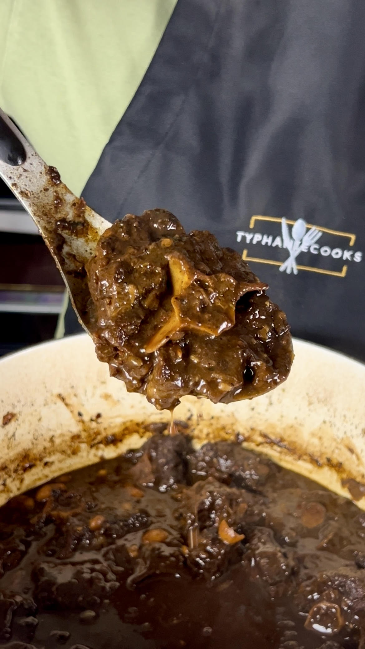 A spoon lifting tender Jamaican oxtail coated in thick, glossy gravy from a pot, showing the rich texture.