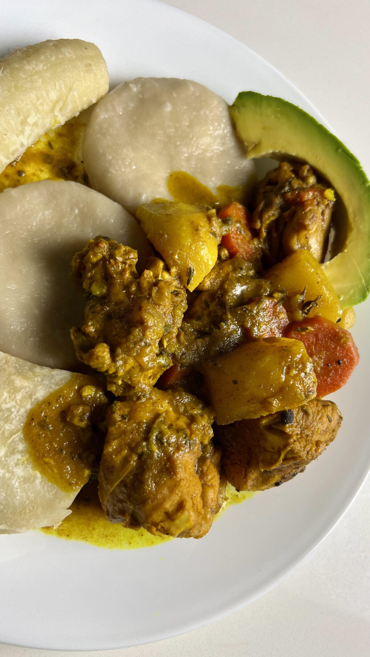 A plated serving of Jamaican curry chicken with potatoes, carrots, dumplings, and avocado on a white plate.