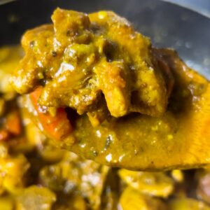 A close-up of a spoon lifting tender Jamaican curry chicken coated in a rich, golden curry sauce from a pan.