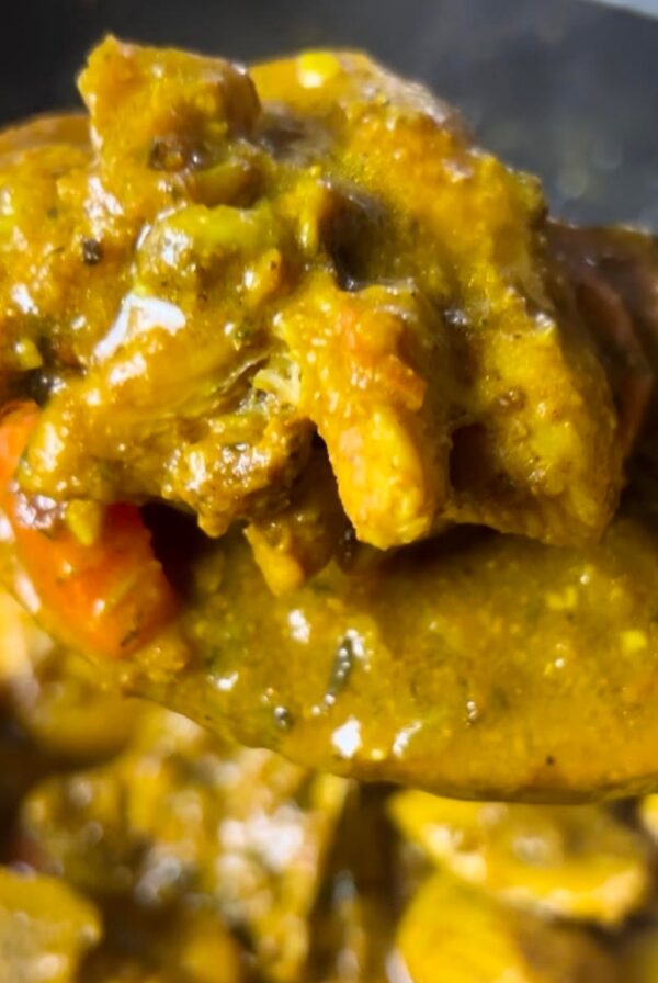 A close-up of a spoon lifting tender Jamaican curry chicken coated in a rich, golden curry sauce from a pan.