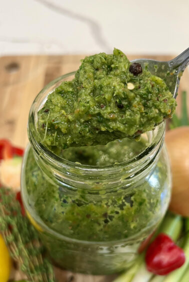 A spoon scooping vibrant Caribbean green seasoning from a jar, highlighting its chunky texture and fresh herb blend.