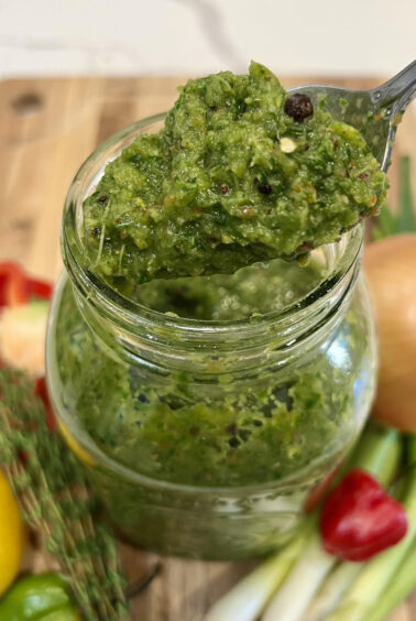 A spoon scooping vibrant Caribbean green seasoning from a jar, highlighting its chunky texture and fresh herb blend.