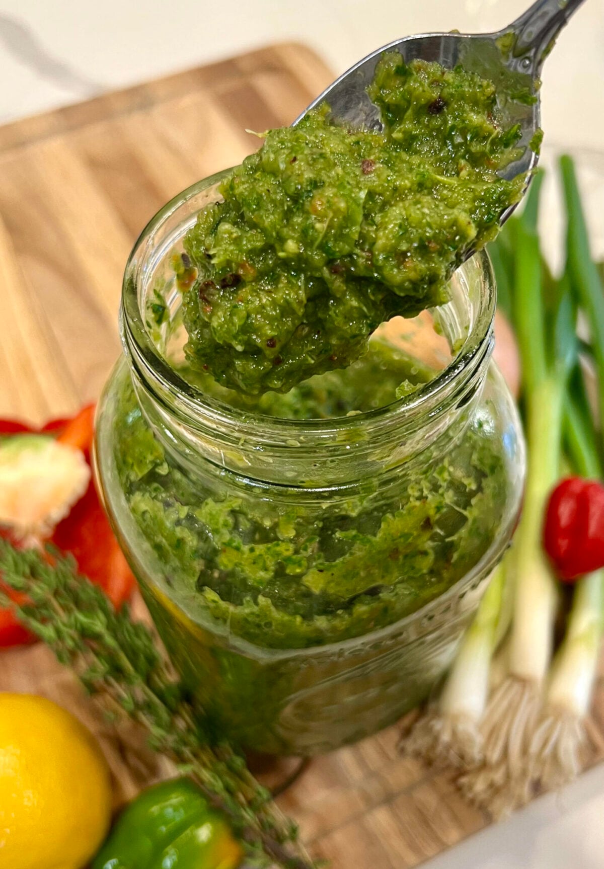 Close-up of a jar filled with homemade green seasoning with visible herbs and spices, surrounded by fresh ingredients.