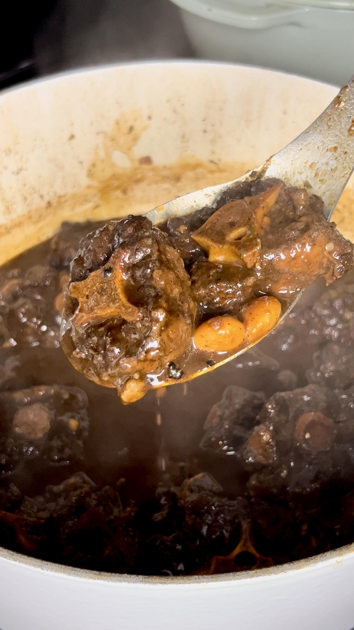 A spoon holding two pieces of tender oxtail and butter beans over a pot of simmering stew with visible steam.