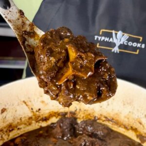 Close-up of a spoonful of braised oxtail with deep brown gravy held over a pot, with the Typhanie Cooks logo visible in the background.