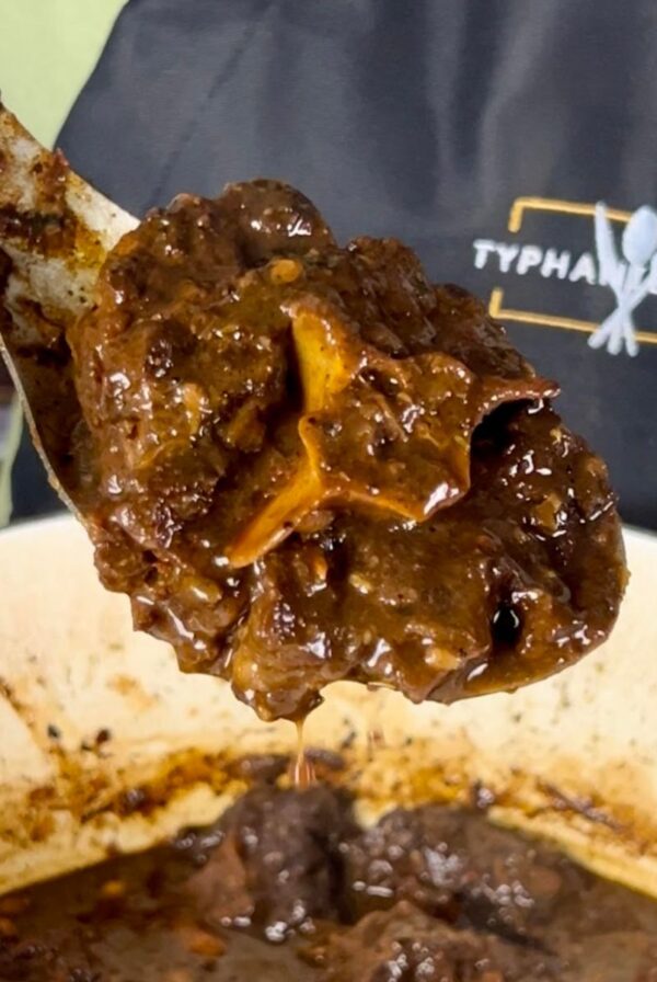 Close-up of a spoonful of braised oxtail with deep brown gravy held over a pot, with the Typhanie Cooks logo visible in the background.