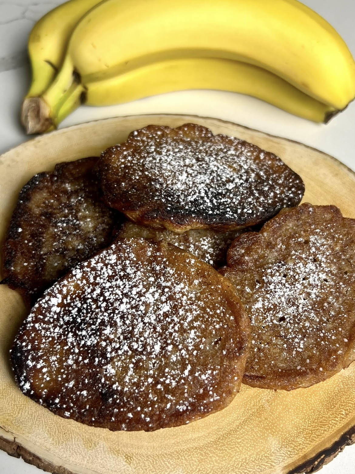 Golden brown banana fritters topped with powdered sugar and arranged on a rustic wooden serving plate with fresh bananas behind them.