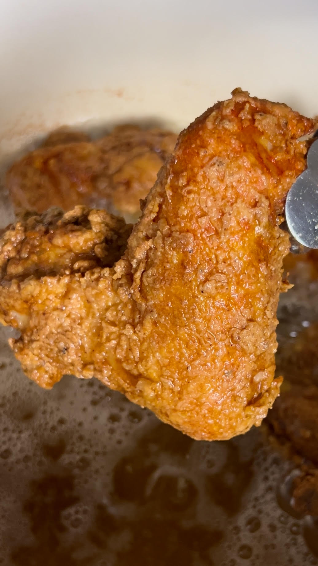 Close-up of a whole fried chicken wing being lifted from hot oil with tongs, showing a textured, crispy exterior.