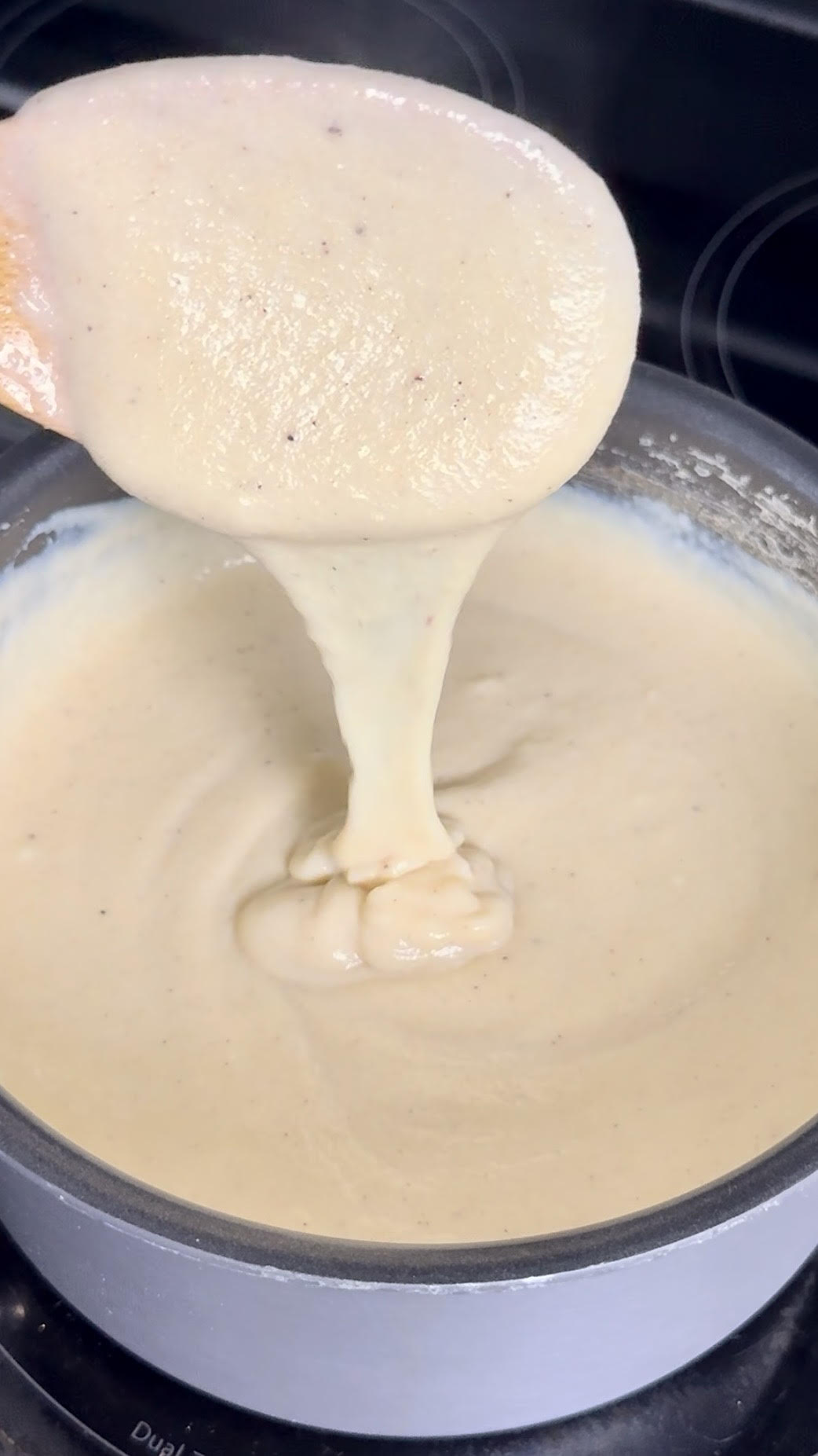 A spoon lifting thick, smooth cornmeal porridge from a pot on the stove, showing its creamy consistency as it stretches back into the pan.
