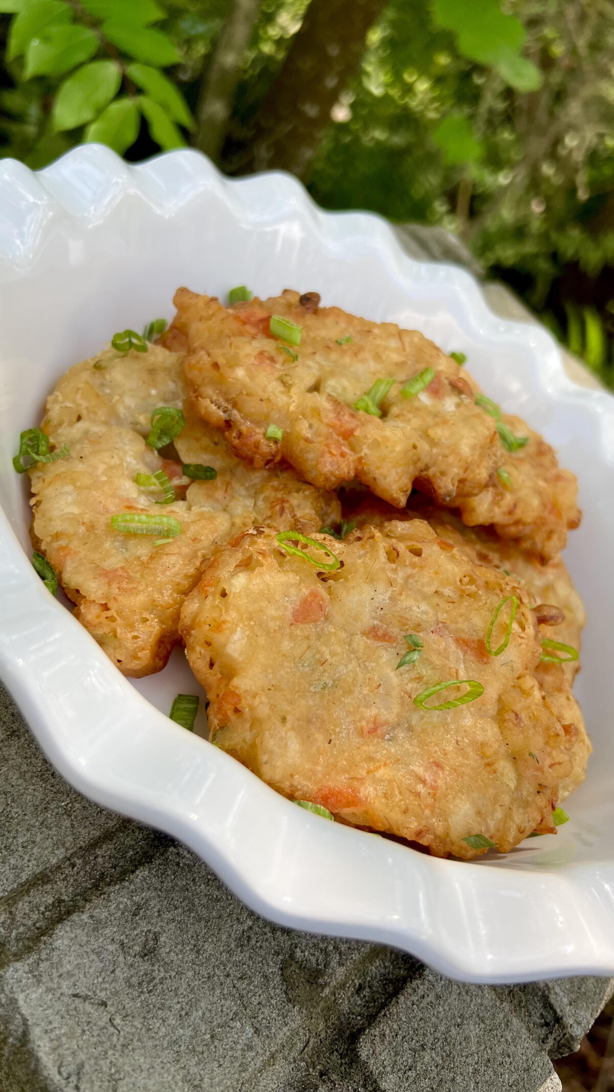 A bowl of saltfish fritters placed outdoors with greenery in the background, showing their lightly crisp edges and soft interior.