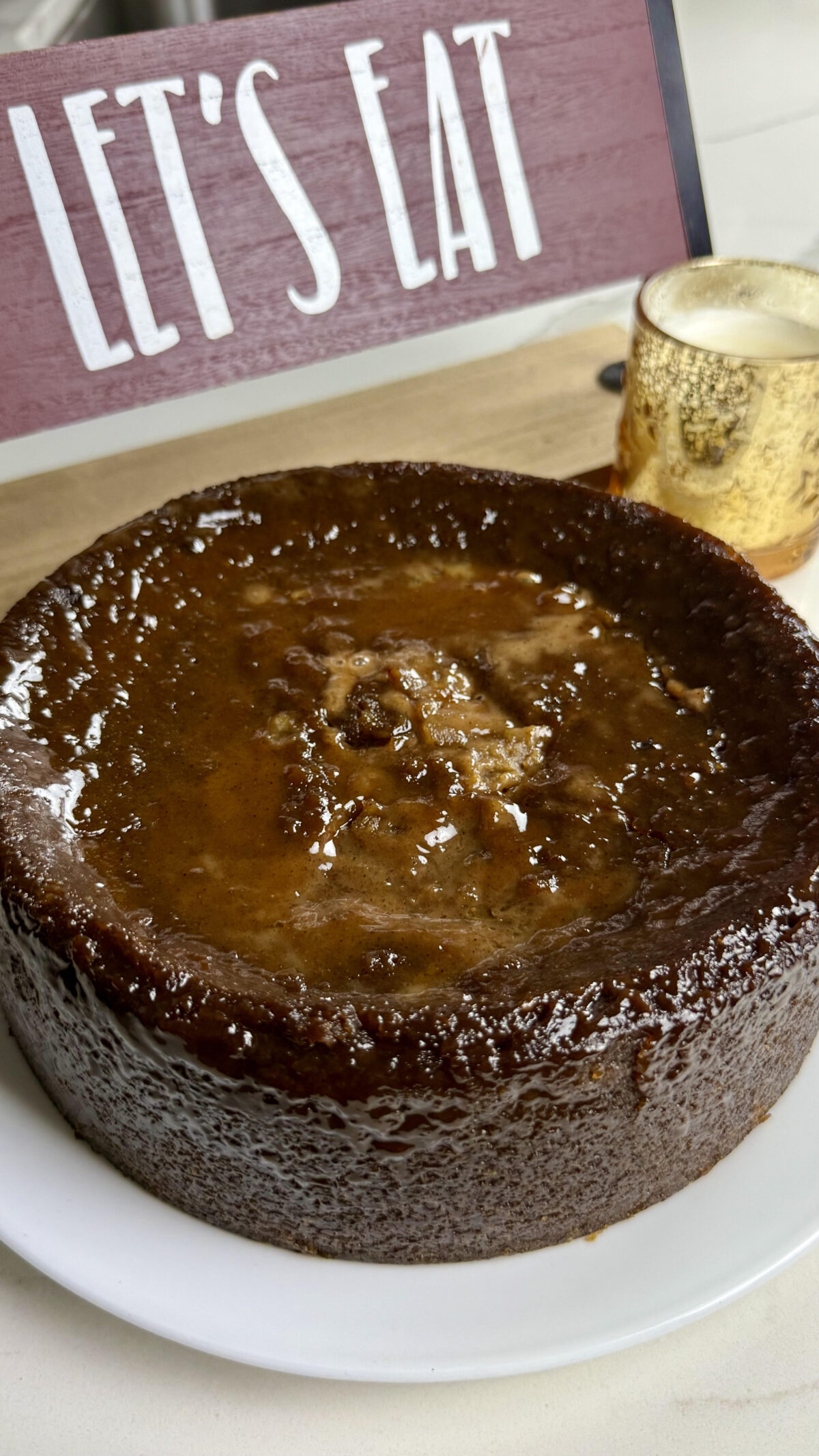 A full round sweet potato pudding with a dark, glossy top sitting on a plate with a &ldquo;Let&rsquo;s Eat&rdquo; sign in the background.