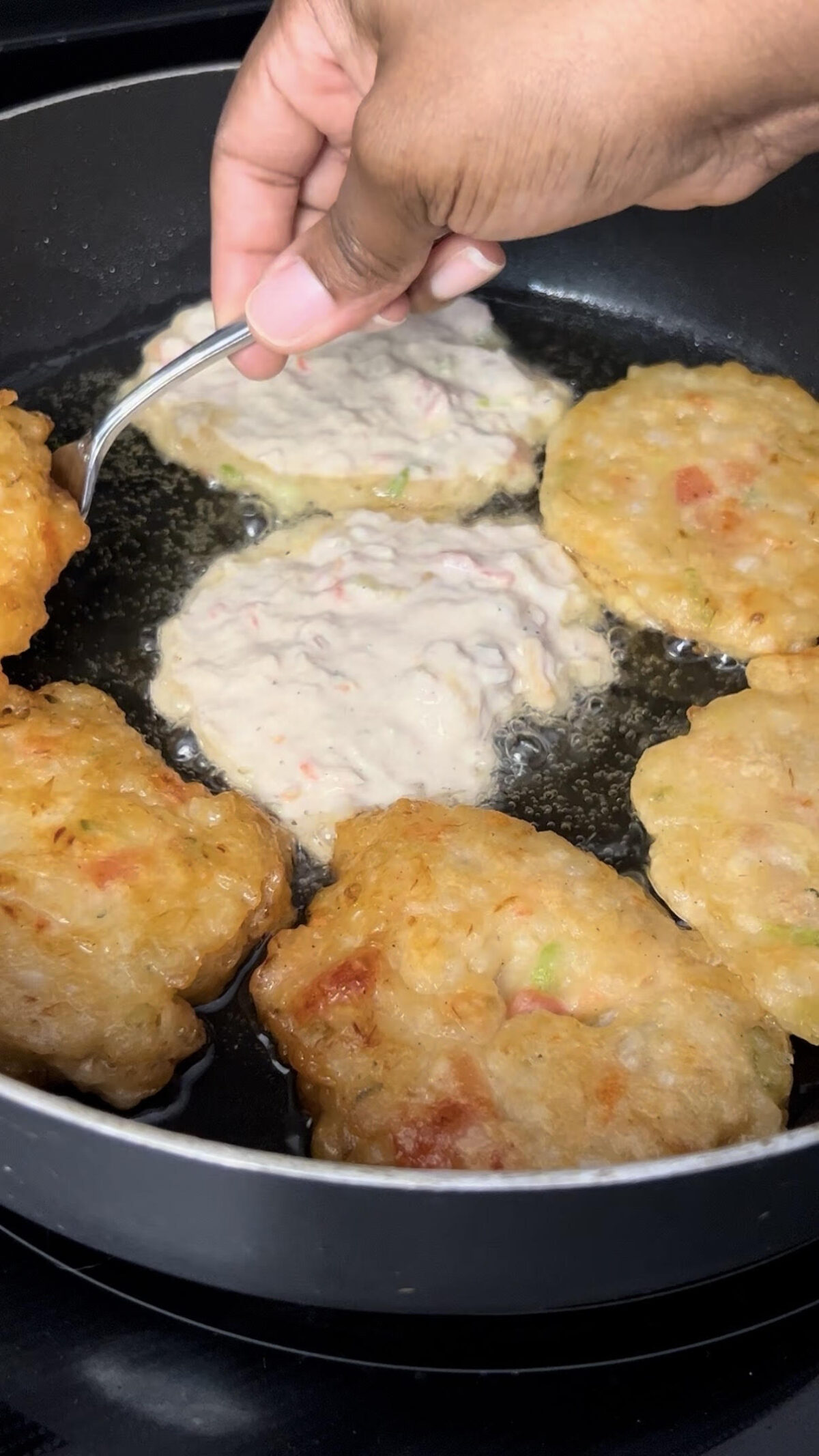 Saltfish fritters frying in hot oil in a skillet as a spoon drops fresh batter into the pan.