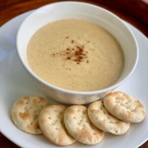 A bowl of creamy Jamaican cornmeal porridge topped with a sprinkle of spice, served on a white plate with round water crackers on the side.
