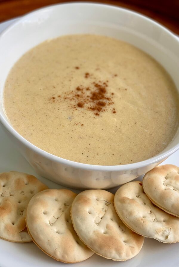 A bowl of creamy Jamaican cornmeal porridge topped with a sprinkle of spice, served on a white plate with round water crackers on the side.