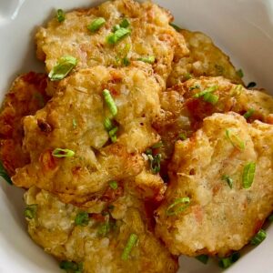 A close-up of golden saltfish fritters stacked in a bowl and garnished with chopped scallions.