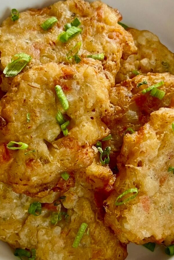 A close-up of golden saltfish fritters stacked in a bowl and garnished with chopped scallions.