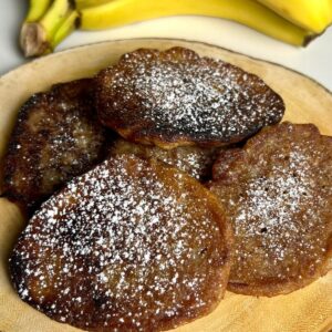 Jamaican banana fritters dusted with powdered sugar and served on a wooden plate with ripe bananas in the background.