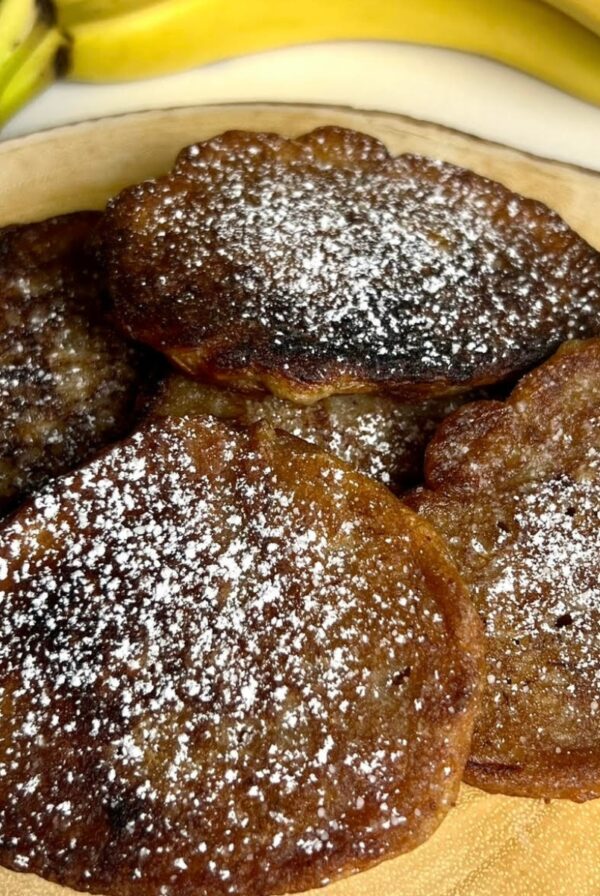 Jamaican banana fritters dusted with powdered sugar and served on a wooden plate with ripe bananas in the background.