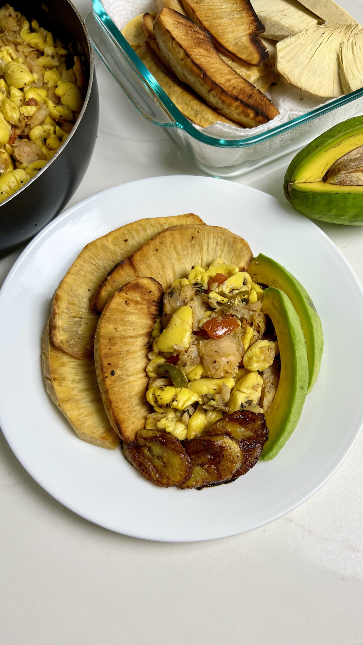 Plate of Jamaican ackee and saltfish with roasted breadfruit, fried sweet plantains, and avocado, with additional breadfruit and ackee visible in the background.