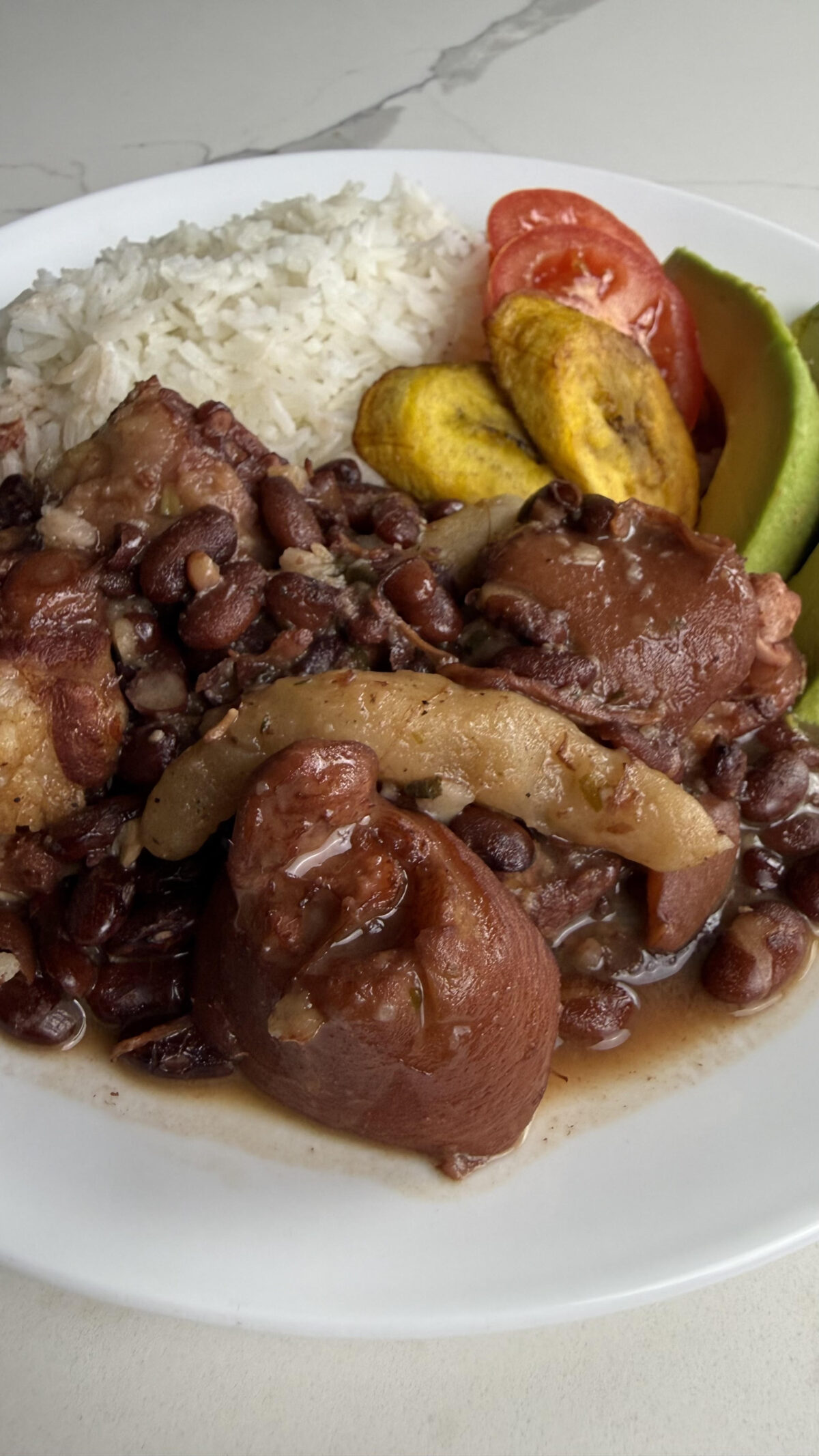 Plate of Jamaican stew peas served with white rice, fried plantains, fresh tomato, and avocado slices.