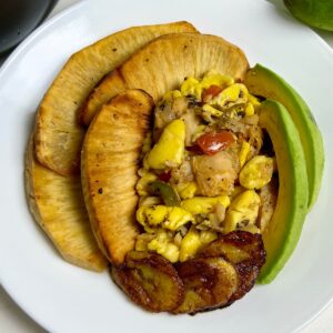 Ackee and saltfish served with roasted breadfruit, fried sweet plantains, and fresh avocado slices on a white plate.