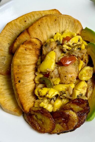 Ackee and saltfish served with roasted breadfruit, fried sweet plantains, and fresh avocado slices on a white plate.