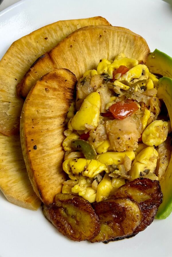 Ackee and saltfish served with roasted breadfruit, fried sweet plantains, and fresh avocado slices on a white plate.