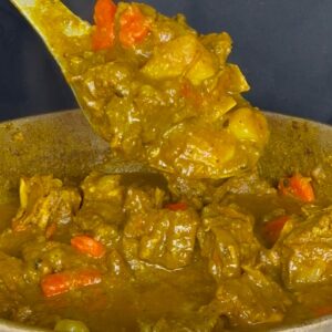 Spoon lifting tender Jamaican curry goat with potatoes and carrots from a pot of rich curry gravy.