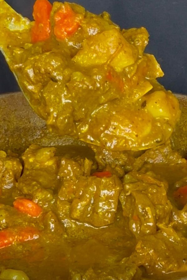 Spoon lifting tender Jamaican curry goat with potatoes and carrots from a pot of rich curry gravy.