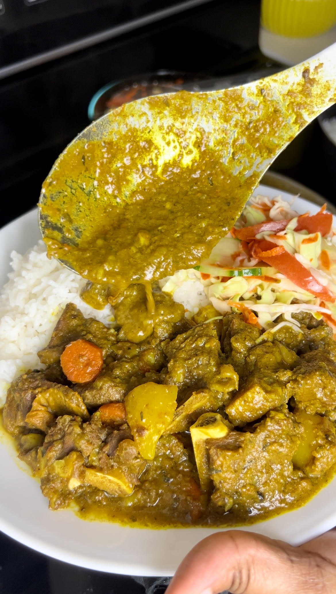 Spoon holding curry sauce over a plate of Jamaican curry goat, potatoes, carrots, rice, and cabbage.