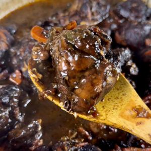 A spoon lifting deep brown Jamaican Brown Stew Chicken out of a pan.
