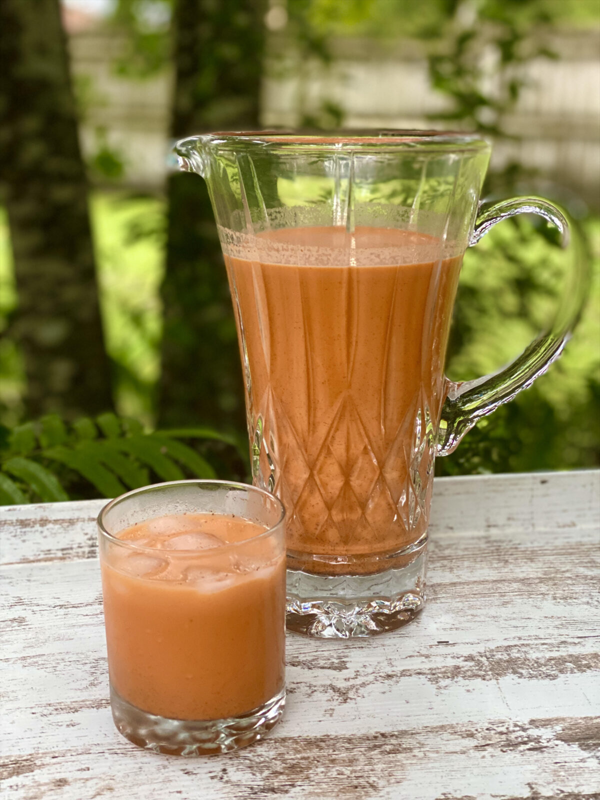 A glass of Jamaican carrot juice with ice sits in front of a glass pitcher filled with carrot juice on an outdoor table.