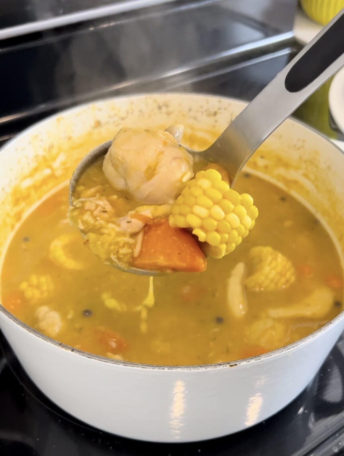 A steaming pot of Jamaican chicken soup with a ladle full of chicken, corn, carrot, and dumplings.