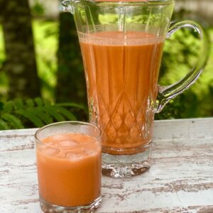 A glass of Jamaican carrot juice with ice sits in front of a glass pitcher filled with carrot juice on an outdoor table.