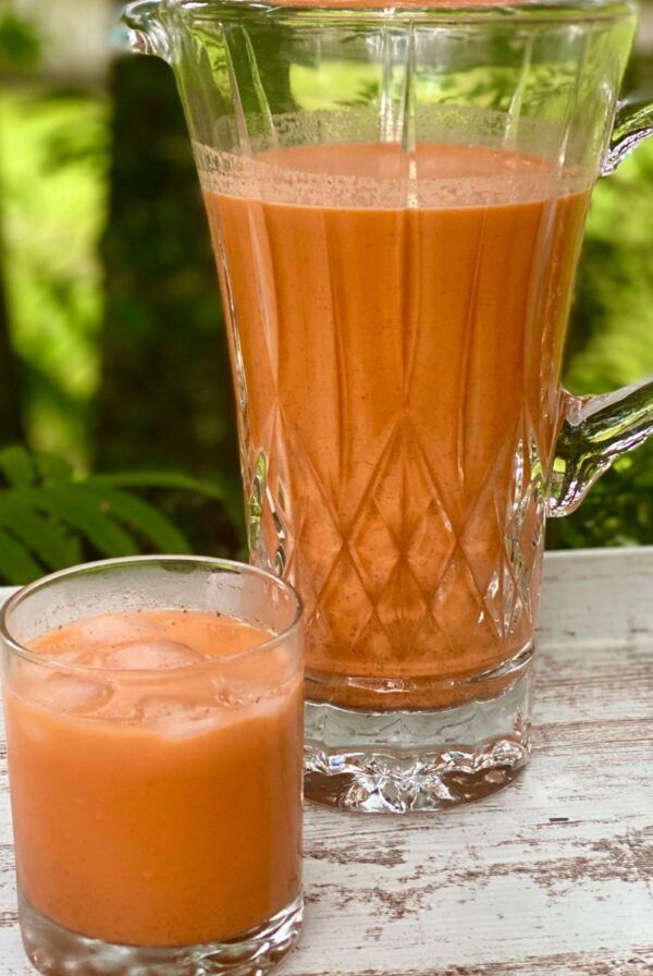 A glass of Jamaican carrot juice with ice sits in front of a glass pitcher filled with carrot juice on an outdoor table.