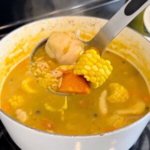 A ladle holds chicken, corn, carrot, and a dumpling over a pot of Jamaican chicken soup.