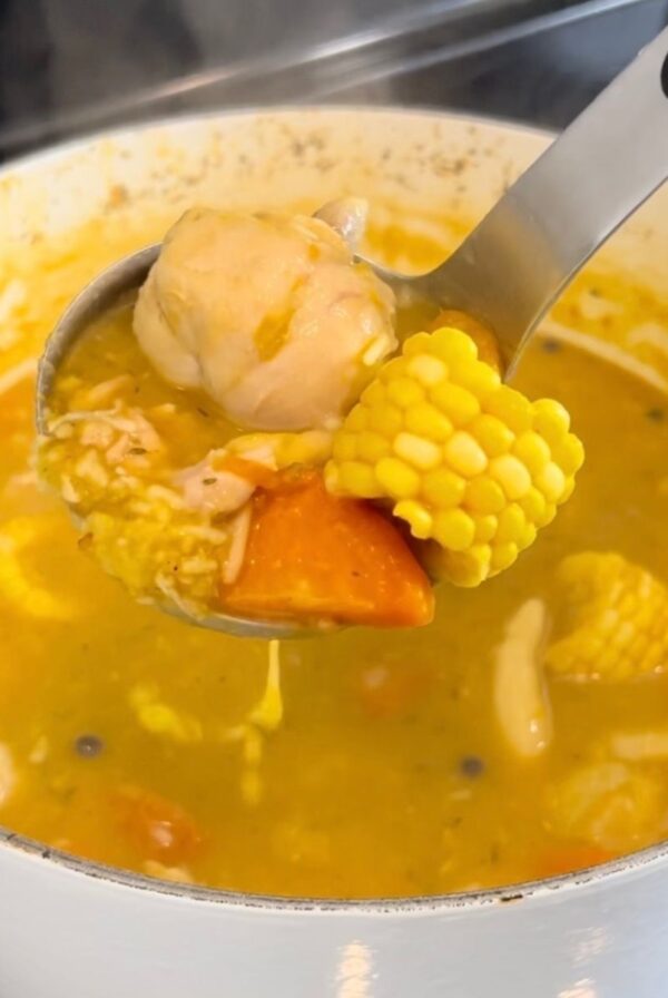 A ladle holds chicken, corn, carrot, and a dumpling over a pot of Jamaican chicken soup.