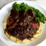Braised beef and carrots in a thick brown gravy are served over mashed potatoes with steamed broccolini in a white bowl.