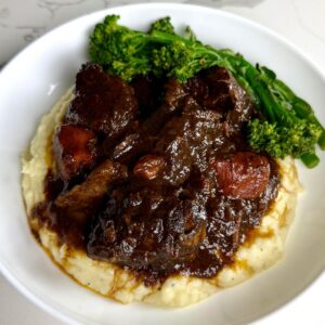 Braised beef and carrots in a thick brown gravy are served over mashed potatoes with steamed broccolini in a white bowl.