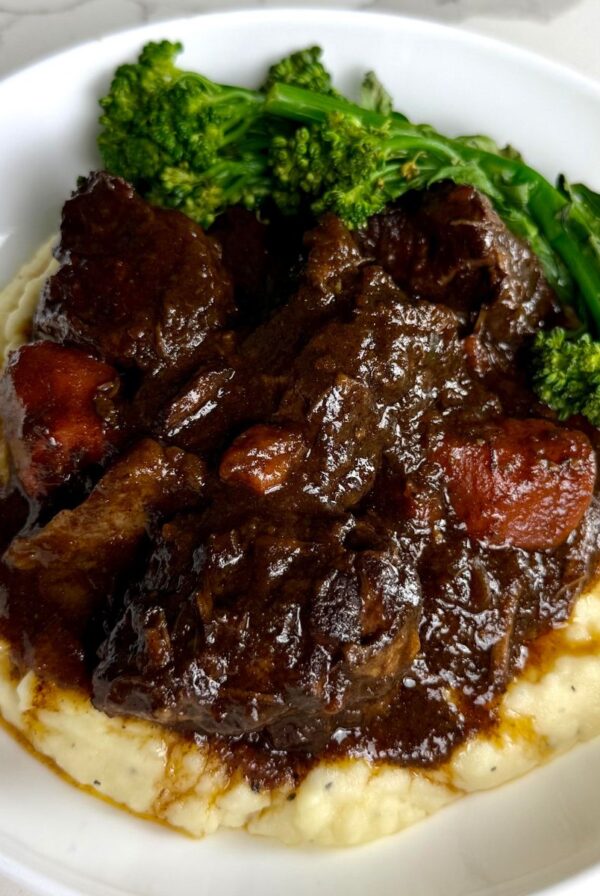 Braised beef and carrots in a thick brown gravy are served over mashed potatoes with steamed broccolini in a white bowl.