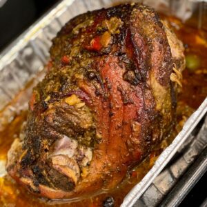 A close-up of a seasoned pork shoulder roast with browned skin, herbs, peppers, and pan juices in a foil roasting pan.
