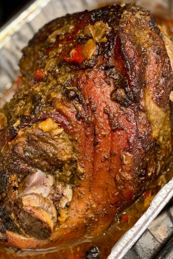 A close-up of a seasoned pork shoulder roast with browned skin, herbs, peppers, and pan juices in a foil roasting pan.