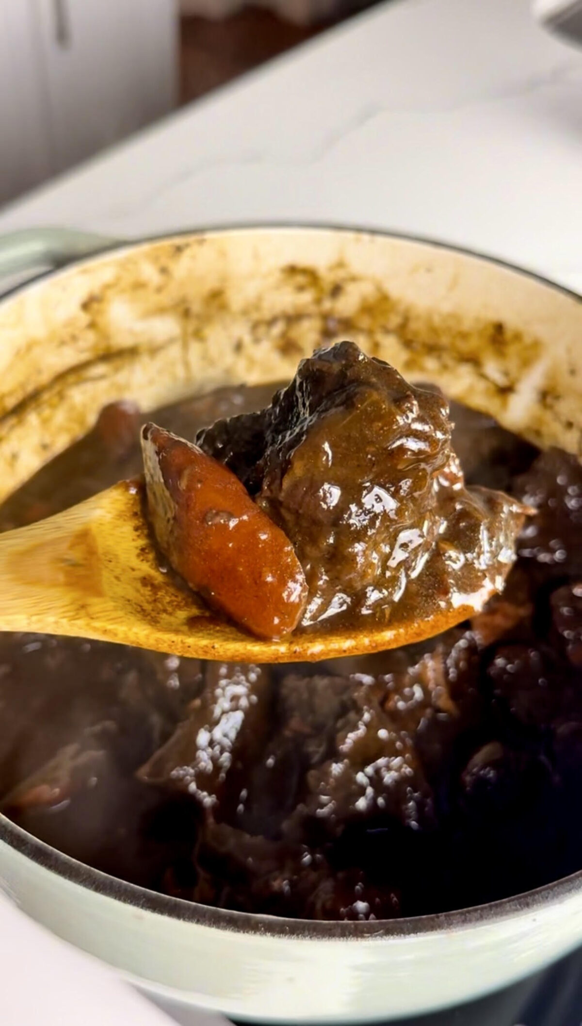 Tender braised beef and carrots coated in a rich brown gravy are lifted from a Dutch oven with a wooden spoon.