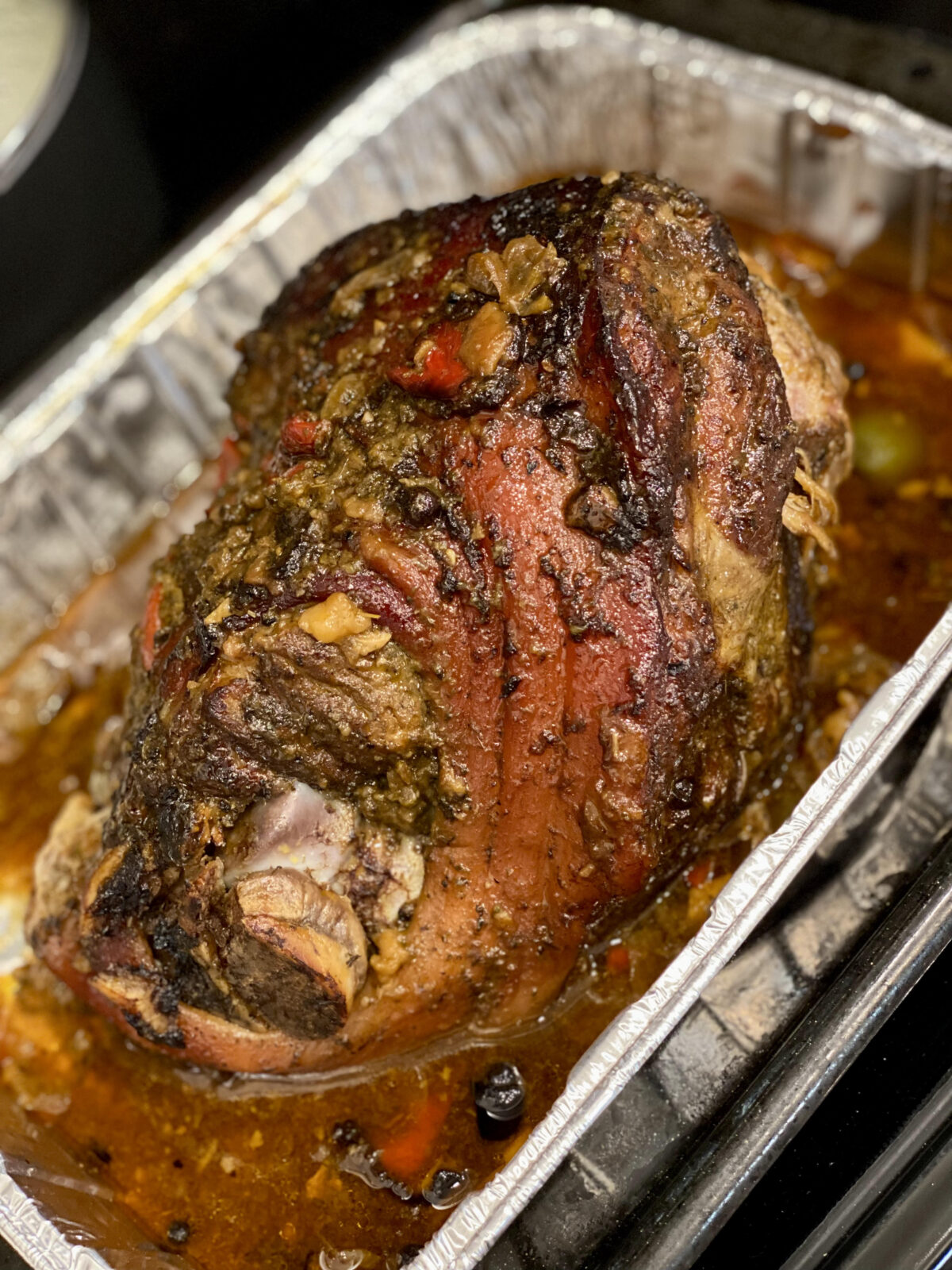 A whole roasted pork shoulder with browned skin, herbs, peppers, and cooking juices in a foil roasting pan.